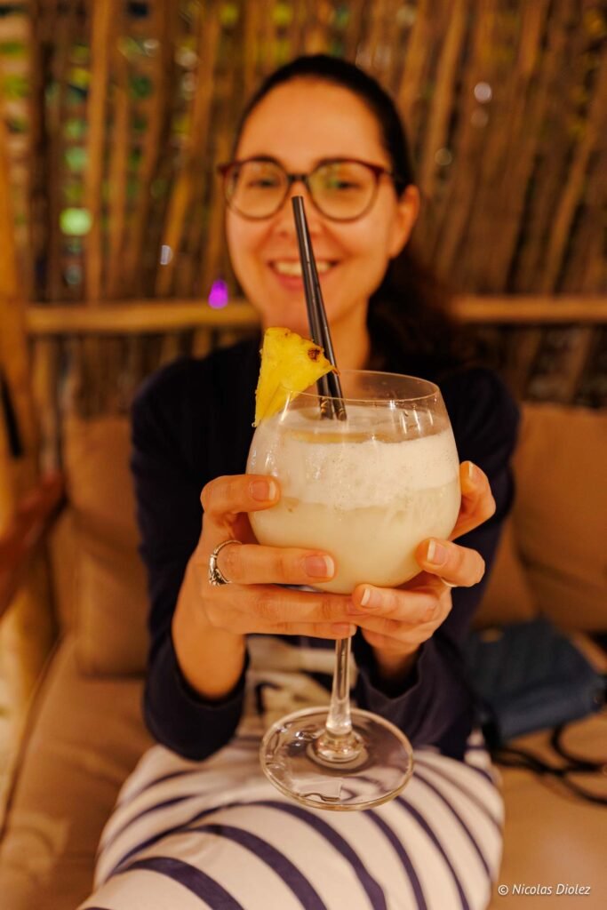 Femme souriante tenant un grand verre de cocktail garni d'ananas lors d'une soirée à l'hôtel Hommage Saint-Martin.