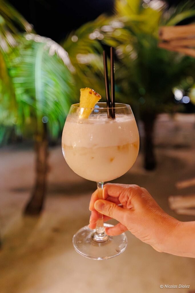 Main tenant un verre de cocktail crémeux garni d'ananas lors d'une soirée à l'hôtel Hommage Saint Martin.