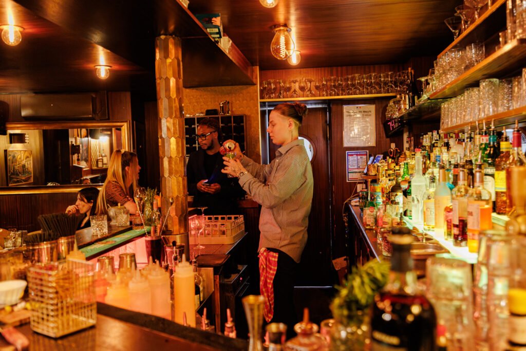 Bartender bereitet in einer gemütlichen Bar einen Cocktail zu, mit Gästen am Tresen im Hintergrund.