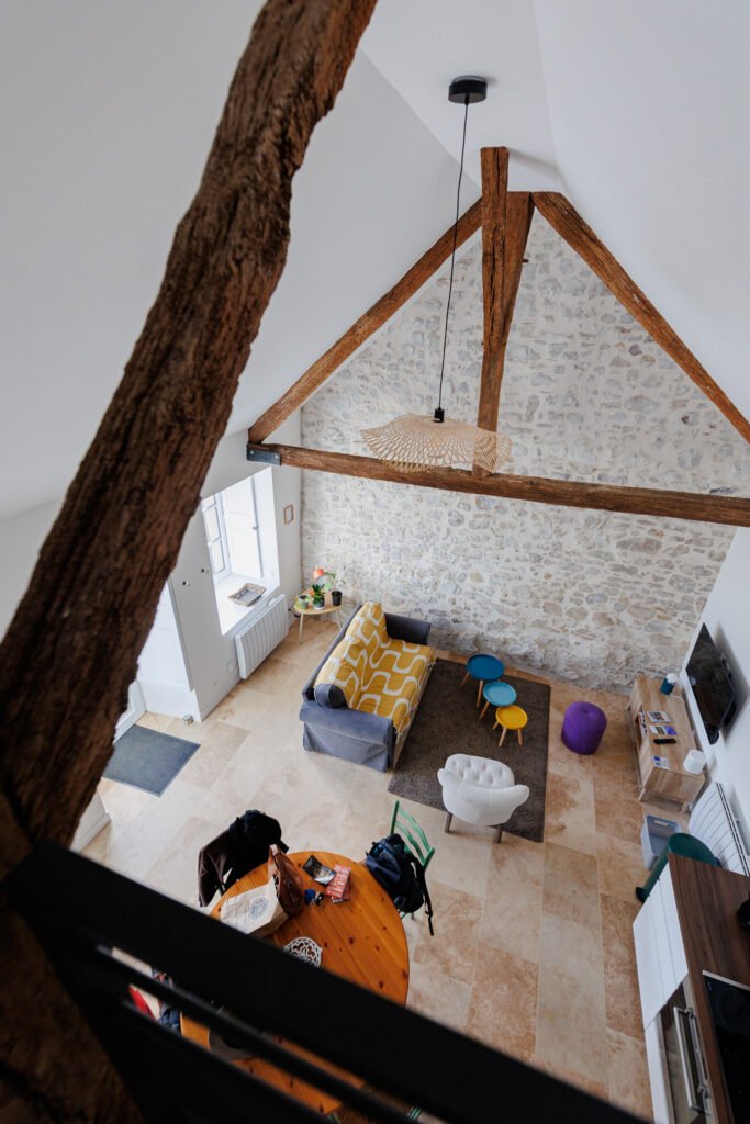 High-angle view of the bright living room with exposed beams and stone wall at Le Logis du Pressoir in Sainte-Suzanne.