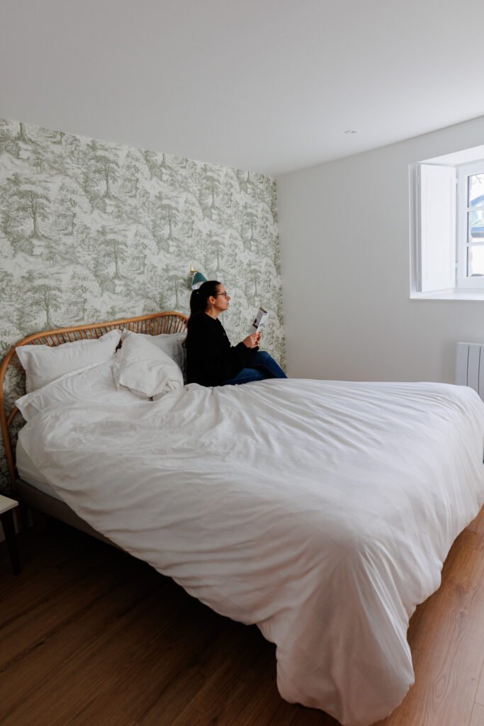 Woman sitting on a comfortable double bed in a bright bedroom at Le Logis du Pressoir gîte in Sainte-Suzanne.