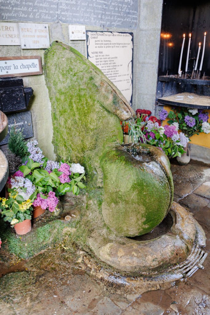 Mossy and flower-adorned stone fountain at the Saint-Céneré oratory, with ex-votos and lit candles in the background.