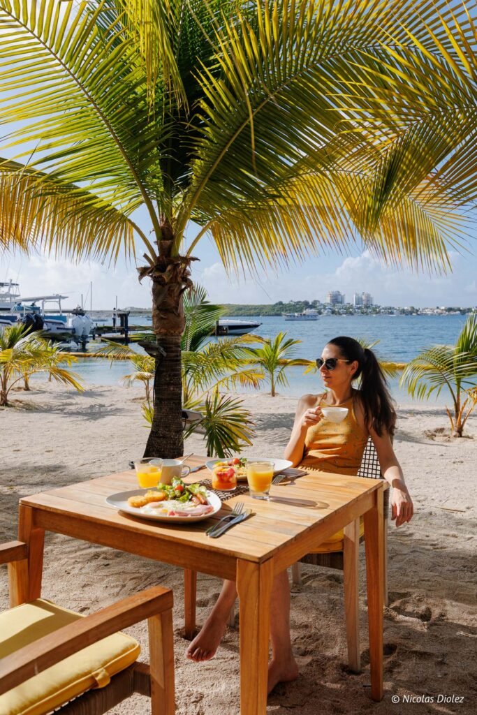 Femme prenant son petit-déjeuner sur une plage tropicale face à la mer, sous un palmier à Saint-Martin.