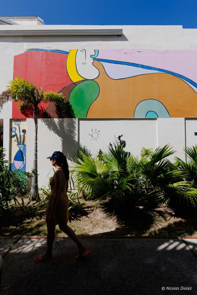 Femme marchant devant une grande fresque murale colorée et des palmiers lors de l'évènement Hommage Saint-Martin.