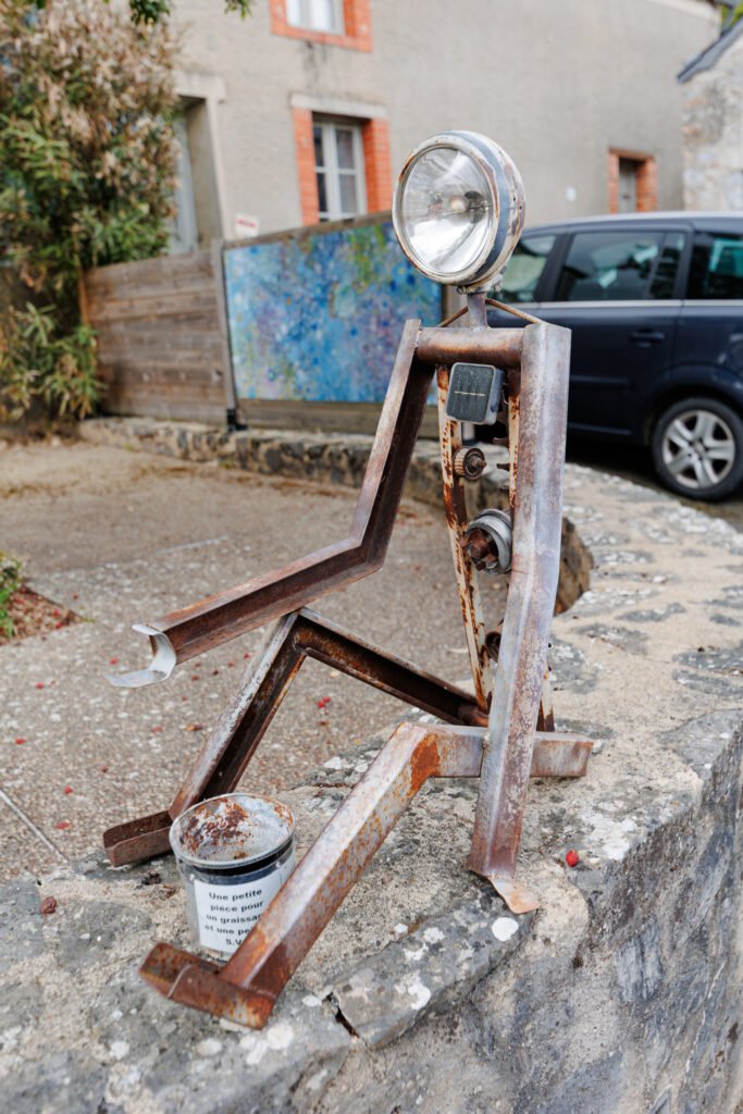 Recycled metal sculpture representing a character sitting on a low stone wall in Saint-Pierre-sur-Erve.