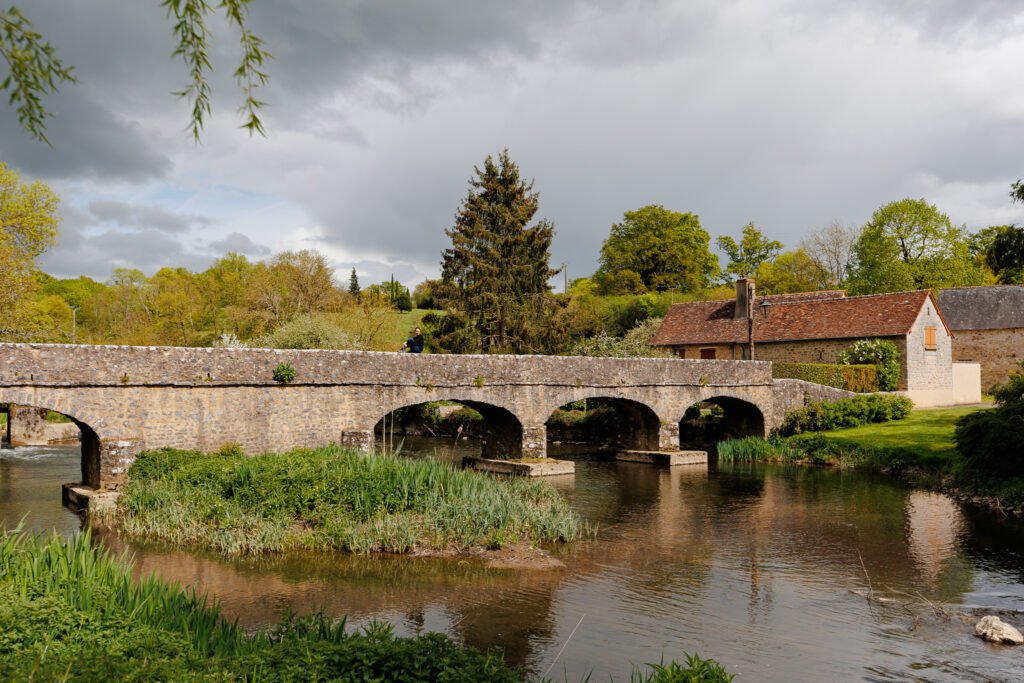 Old stone bridge spanning the Erve river in Saint-Pierre-sur-Erve under a cloudy sky.