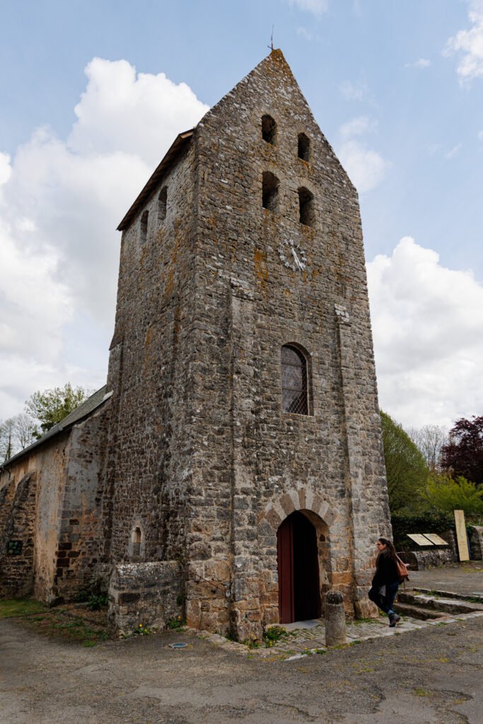 Stone facade and bell tower of the medieval church of Saint-Pierre-sur-Erve with a person walking past.