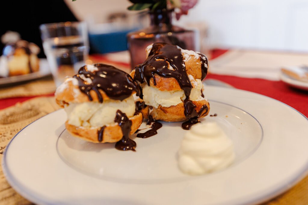 Two cream puffs topped with hot chocolate and served with whipped cream on a plate.